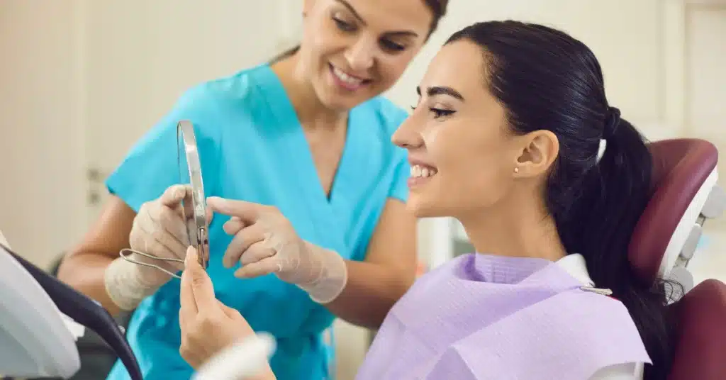A woman in a dental chair smiling into a handheld mirror while a dental assistant in blue scrubs explains her results during a checkup.