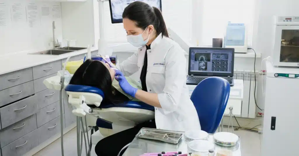 Female dentist wearing gloves and a mask performing a dental procedure on a patient in a modern dental office, with dental X-ray images displayed on a nearby computer screen.