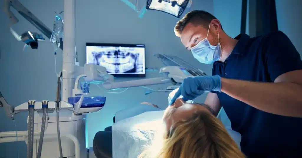 Male dentist in surgical mask examining a patient with dental X-ray visible on monitor in modern dental clinic