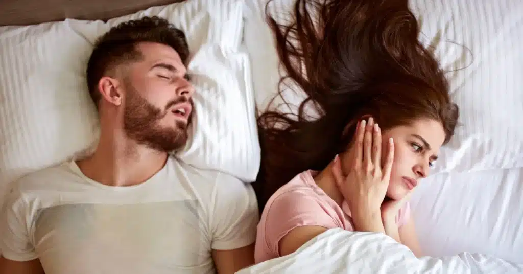 A frustrated woman covering her ears while lying next to a man snoring loudly, illustrating the impact of snoring or sleep apnea on couples.