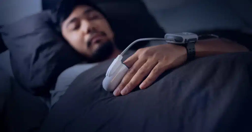 A man sleeping in bed wearing a sleep study sensor on his finger and wrist to monitor sleep apnea or other sleep disorders overnight.