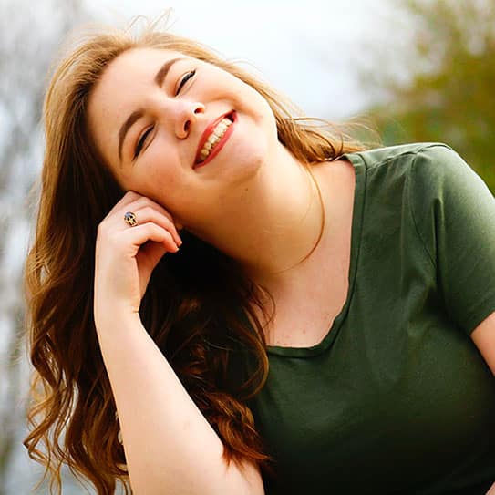 A young woman in a green shirt smiling brightly while enjoying an outdoor setting.