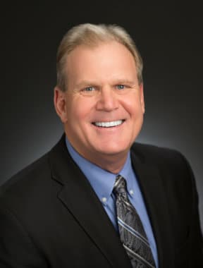 A professional headshot of Dr. Gregg C. Hendrickson, smiling in a suit and tie against a dark background.