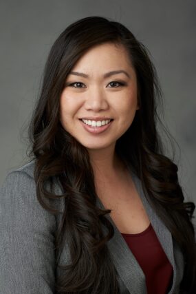 A professional headshot of Dr. Oanh Le, smiling in a suit and tie against a gray background.