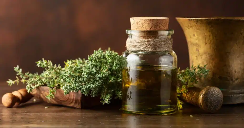 A glass jar of thyme essential oil with a cork lid, surrounded by fresh thyme sprigs, a wooden scoop, and a mortar and pestle on a rustic table.