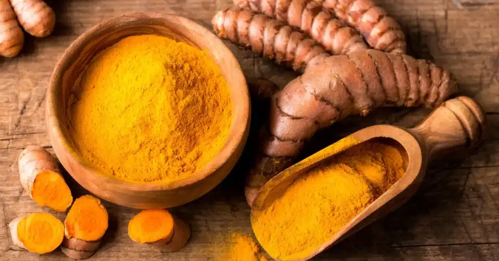 A wooden bowl and scoop filled with bright yellow turmeric powder surrounded by fresh turmeric roots on a rustic wooden surface.