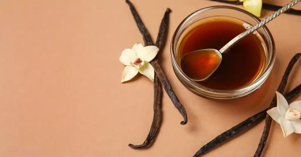 A small glass bowl filled with vanilla extract and a spoon, surrounded by vanilla beans and white vanilla flowers on a brown background.
