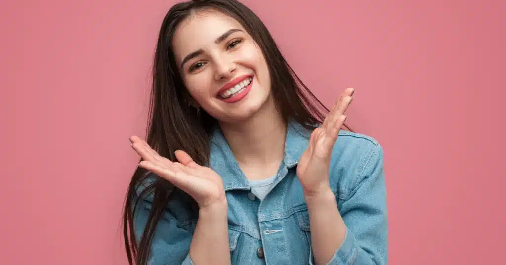 Happy young woman with long dark hair smiling confidently, showing straight white teeth against a pink background