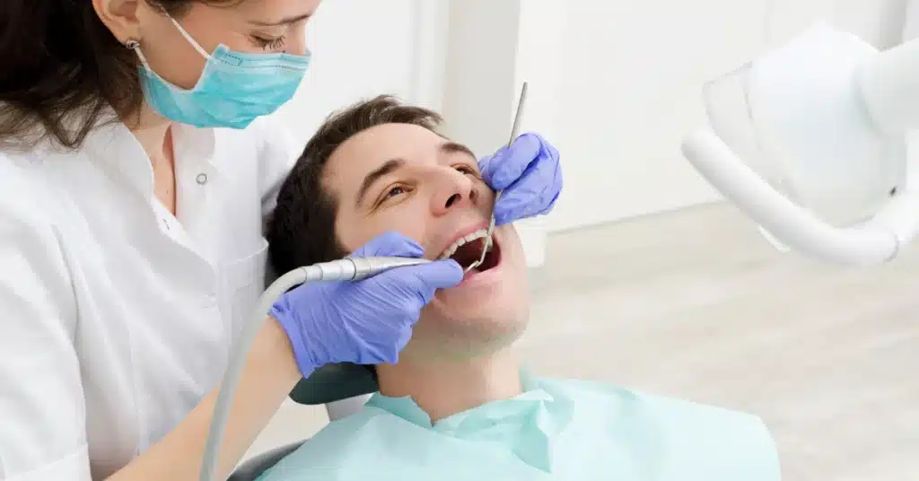 Dental hygienist in gloves and mask performing a routine teeth cleaning on a smiling male patient in a modern dental clinic